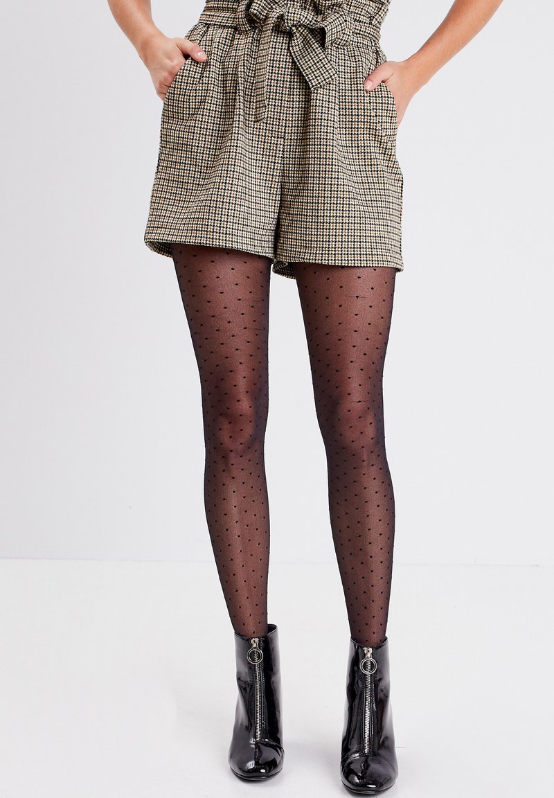 Shorts à carreaux en beige et noir, taille haute avec une ceinture en tissu, associés à des collants noirs transparents à pois et des bottes noires brillantes à la cheville.