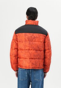 Doudoune avec un motif floral orange, panneau d'épaule en nylon noir et col haut. Design texturé et rembourré avec des poignets élastiques.