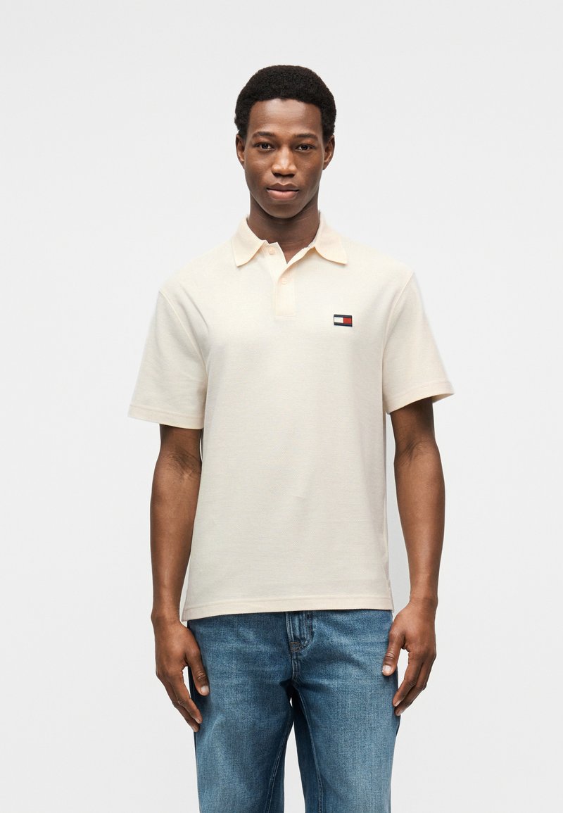 Μπεζ κοντομάνικο πόλο πουκάμισο από υφασμάτινη υφή, με μικρό πολύχρωμο λογότυπο στο αριστερό στήθος. Συνδυασμένο με μπλε τζιν.