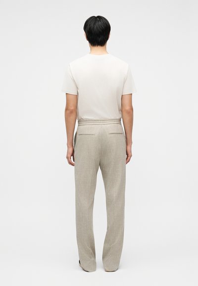 Άνδρας με κοντά μαύρα μαλλιά, φορώντας λευκό κοντομάνικο πουκάμισο και μπεζ καρό παντελόνι, που στέκεται γυρισμένος προς τα πίσω σε απλό φόντο.