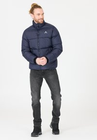 Veste matelassée marine avec un col haut, une fermeture éclair à l’avant et un logo de marque. Assortie à un jean foncé et des bottes de randonnée noires. Texture lisse et rembourrée.