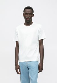 Giovane uomo con capelli corti, vestito con una maglietta bianca semplice e jeans azzurri chiari, in piedi davanti a uno sfondo bianco.