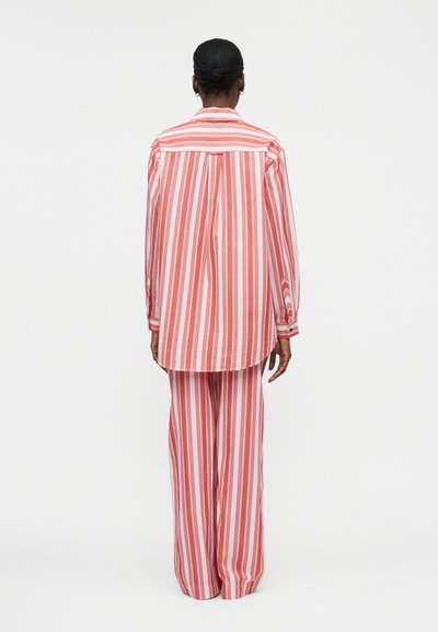 Isik seisab seljaga, seljas lahtine punase ja valge vertikaaltriibuline särk ning sobivad laia säärega püksid tavalise taustaga.