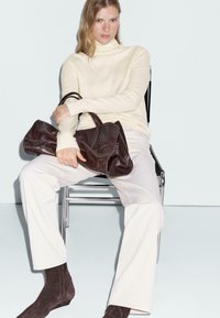 Brązowa skórzana torebka o teksturowanej powierzchni, noszona na jasnej kremowej swetrze z golfem i białych spodniach, siedząca na metalowym krześle.