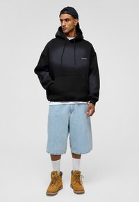 Zwarte en grijze gradient hoodie met trekkoorden, grote voorkantzak, denim shorts en gele werklaarzen. Witte sokken zichtbaar.