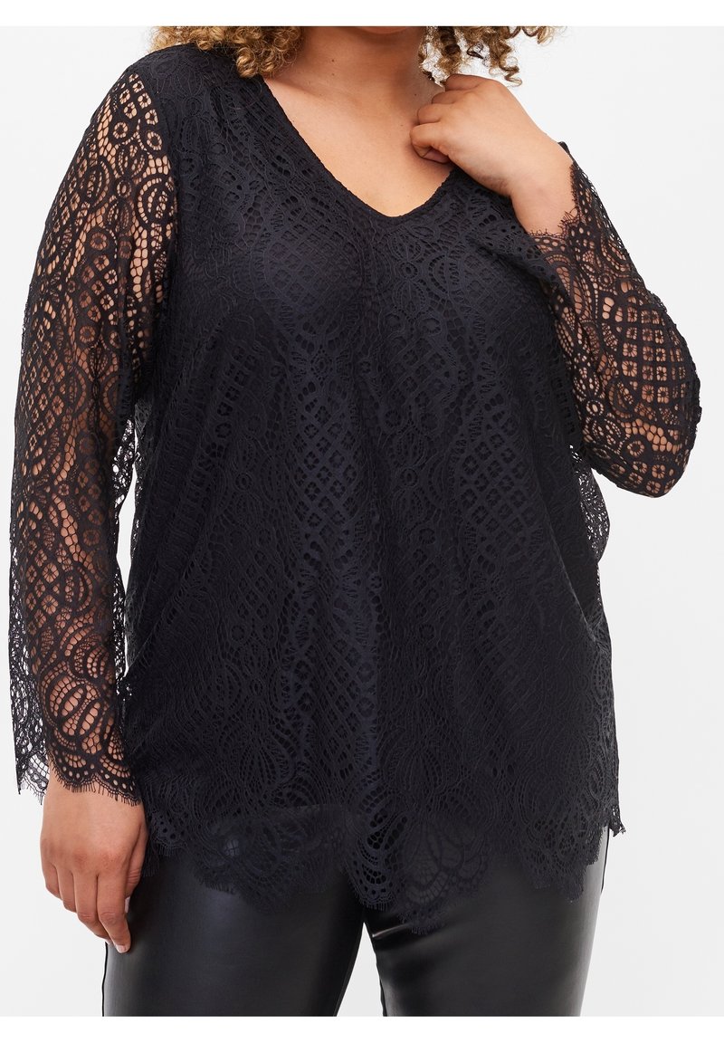Femme portant un top en dentelle noire à manches longues avec un décolleté en V et des bords festonnés, associé à un pantalon en cuir noir sur fond blanc.