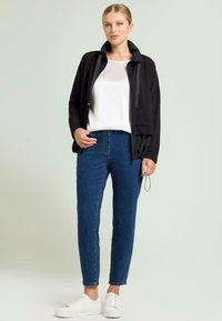 Chaqueta de tela negra con cuello alto y detalle de cordón; combinada con una camiseta blanca y vaqueros azules; usada con zapatillas blancas.