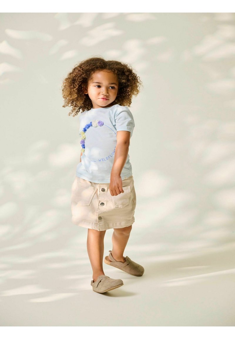 Jeune fille aux cheveux bouclés portant un t-shirt bleu clair avec un motif floral, une jupe beige boutonnée et des chaussures beiges sans lacet, debout sur un fond clair.