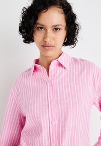 Camisa rosa com riscas, com um colarinho abotoado, apresentando riscas verticais brancas e um tecido leve. Detalhes notáveis incluem botões rosas.