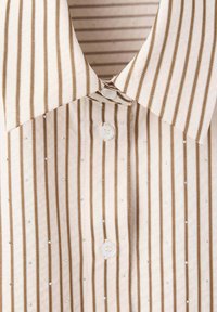 Gros plan d'une chemise beige avec des rayures verticales marron, des boutons blancs et de petits clous argentés dispersés sur le tissu.