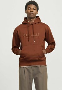 Brun hættejakke med justerbare snore, der har en frontlomme og prægede "ORIGINALS" logo. Båret med lyse bukser.