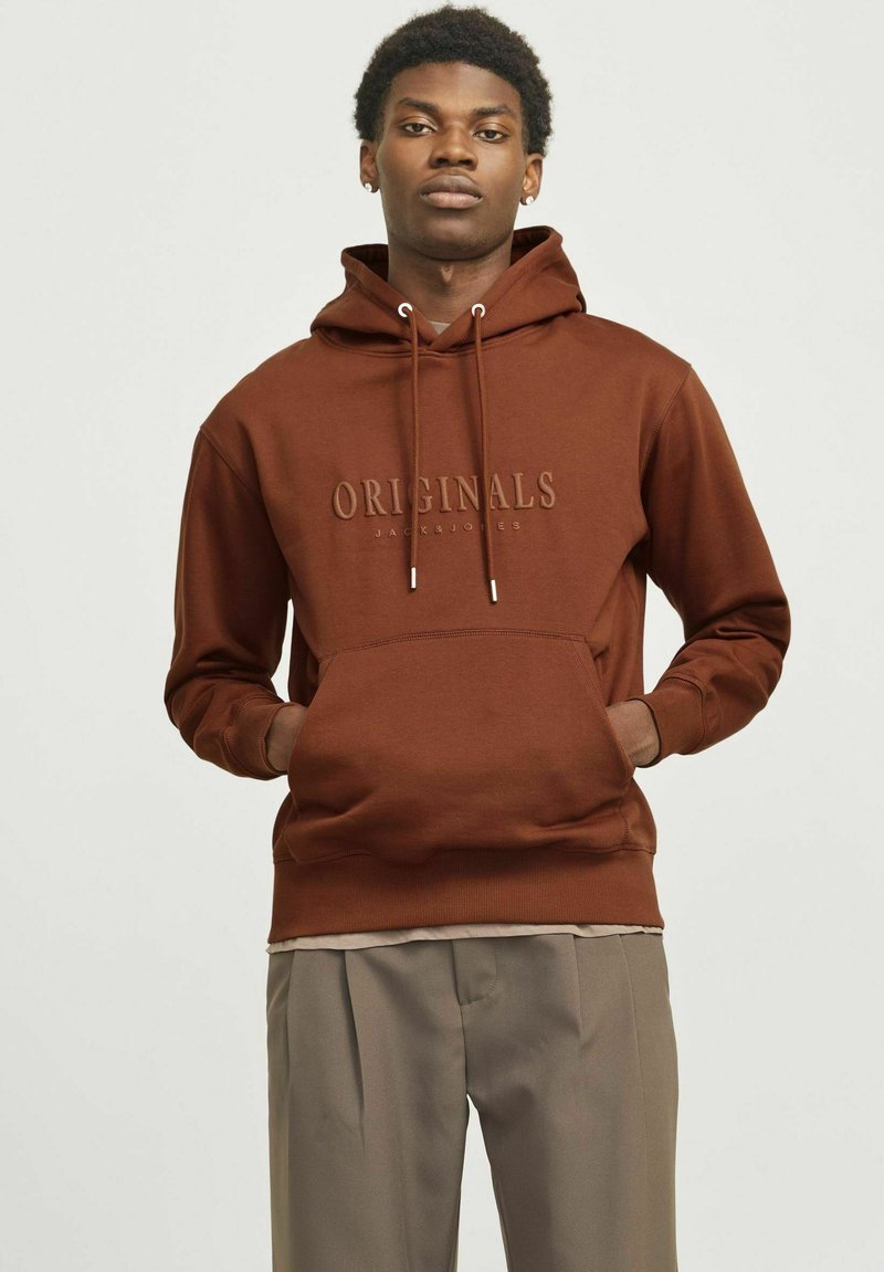 Brun hættejakke med justerbare snore, der har en frontlomme og prægede "ORIGINALS" logo. Båret med lyse bukser.