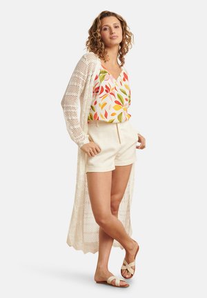 Lichte, crèmekleurige gehaakte cardigan over een kleurrijke, patroon tanktop met bladeren; gecombineerd met beige shorts en platte sandalen.