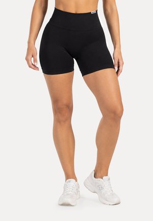 Femme portant un short de sport noir taille haute et des chaussures de course blanches, debout les jambes légèrement écartées sur un fond uni.