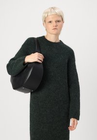 Μαύρη δερμάτινη τσάντα χεριού με στρογγυλή μορφή και έναν μόνο ιμάντα, εκτεθειμένη δίπλα σε ένα σκούρο πράσινο πλεκτό φόρεμα με υφή.