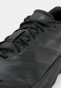 Chaussure de sport noire en matériau synthétique texturé avec un design en maille complexe, dotée de lacets foncés et d'accents de marque gris.