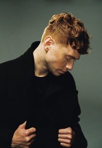 Portret van een persoon met kort, krullend kastanjebruin haar, gekleed in een zwarte jas, handen grijpen de jas vast, hoofd licht gebogen tegen een neutrale achtergrond.