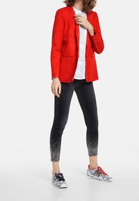 Rød ruskinds blazer, hvid mønstret skjorte, sorte jeans med gradient glimmerdetaljer, og sorte og hvide grafiske sneakers med røde snørebånd.
