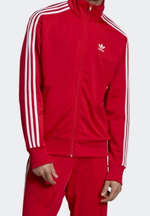 Homme portant un survêtement rouge Adidas avec des bandes blanches le long des manches et des jambes, debout devant un fond uni.