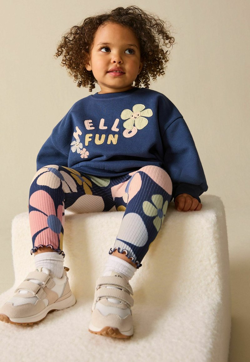 Niño pequeño con cabello rizado sentado sobre un bloque blanco texturizado, vistiendo un suéter azul marino con las palabras "Hola Diversión" y diseños de flores, y leggings a juego con estampado floral.