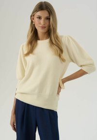 Pull crème léger avec une finition texturée, col rond et manches bouffantes. Porté avec un pantalon bleu marine, avec des liens latéraux sur les manches.
