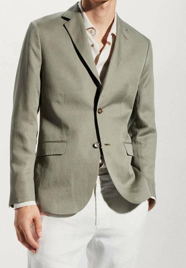 Homme portant un blazer vert clair sur une chemise blanche et un pantalon blanc, avec une main dans la poche sur un fond uni.