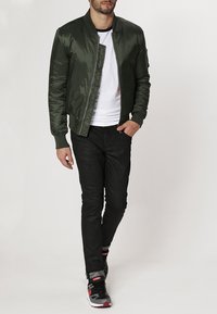 Grön satinbomberjacka med dragkedja framtill, ribbade muddar och en vit t-shirt. Kombineras med svarta byxor och sneakers med röda detaljer.
