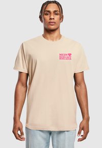 Beige bomulds t-shirt med korte ærmer, der har pink tekst, som siger "MEIN ♥ SCHLÄGT SCHLAGER" på venstre brystområde.