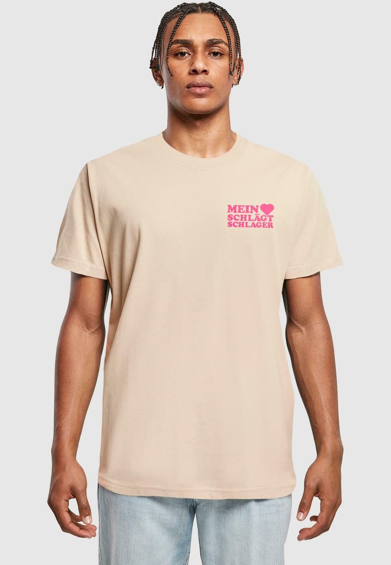 Beige bomulds t-shirt med korte ærmer, der har pink tekst, som siger "MEIN ♥ SCHLÄGT SCHLAGER" på venstre brystområde.