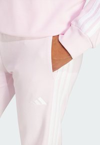 Lys rosa Adidas treningsjakke med hvit tre-stripesdesign og lommeryddig, med myk stoff og ribbestrikkede mansjetter.