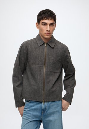 Ung mand iført en grå lynlåstrøje med krave og brystlomme, kombineret med lyseblå jeans, stående mod en hvid baggrund.