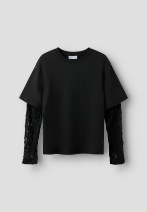 T-shirt noir à manches courtes superposé à de longues manches en dentelle noire, sur un fond gris clair.