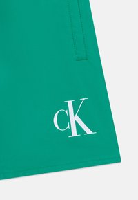 Κοντινό πλάνο πράσινου υφάσματος με ραμμένη τσέπη και λευκό λογότυπο "cK" που έχει τυπωθεί κοντά στο τελείωμα.