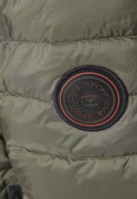 Tela de chaqueta acolchada de color verde oliva con textura acolchada, que presenta un parche redondo negro con acentos rojos y un logo bordado.