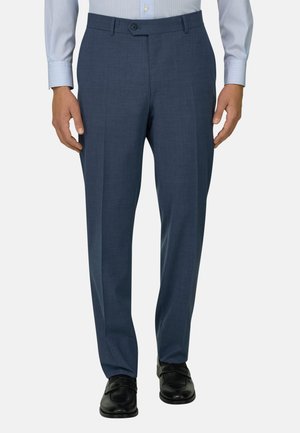 Muž v tmavě modrých kalhotách na míru, světle modré pruhované košili a černých kožených společenských botách, stojící proti jednobarevnému pozadí.