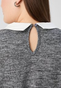 Suéter de punto gris con un cuello blanco, abertura en la parte posterior con forma de llave y cierre de botón. Textura suave con un patrón sutil.