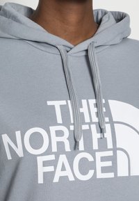Felpa grigia con cappuccio e un grande logo bianco che reca la scritta "THE NORTH FACE." Presenta un cappuccio con cordini e un orlo a coste per una vestibilità aderente.
