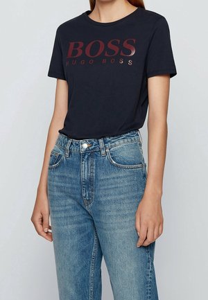 Mustapohjainen puuvillapaita, jossa on kiiltävä burgundin värinen "BOSS HUGO BOSS" -logo. Yhdistetty vaaleansinisiin korkeavyötäröisiin farkkuihin, joissa on haalistunut pinta.