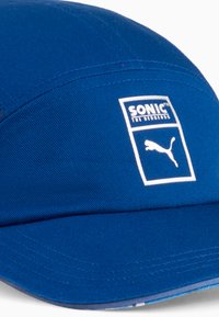 Blå kasket med et struktureret design, der har en hvid grafiklabel med "SONIC THE HEDGEHOG" og Puma-logoet. Glat stofstruktur.