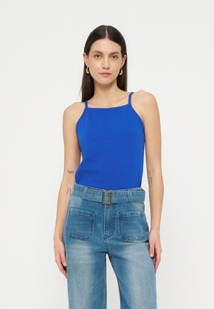 Γυναίκα με μακριά σκούρα μαλλιά, φοράει μπλε φωτεινή αμάνικη μπλούζα, ανοιχτόχρωμα τζιν ψηλόμεσα με ζώνη και χρυσούς κρίκους στα αυτιά.