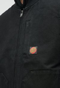 Μαύρο bomber μπουφάν με φερμουάρ, ραμμένο γιακά και κόκκινο και κίτρινο λογότυπο "Santa Cruz" στο αριστερό στήθος.