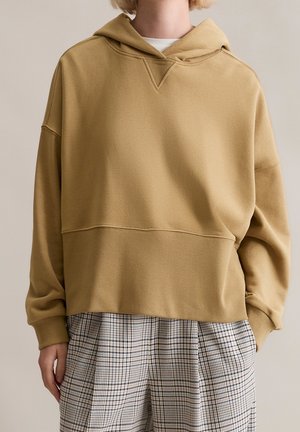 Personne portant un sweat à capuche ample beige associé à un pantalon à carreaux noir et blanc taille haute, debout avec une main dans la poche.