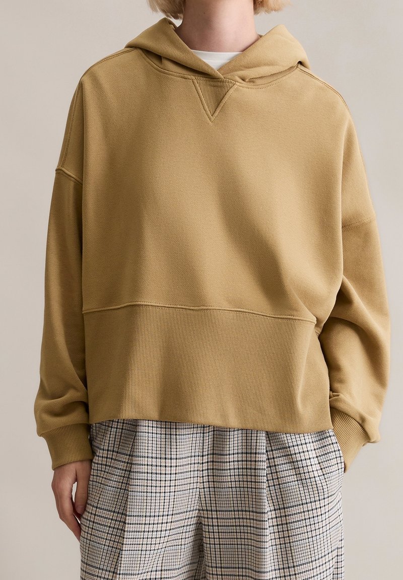 Personne portant un sweat à capuche ample beige associé à un pantalon à carreaux noir et blanc taille haute, debout avec une main dans la poche.