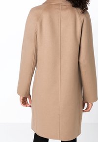Béžový vlněný kabát se vzpřímeným střihem, dlouhými rukávy a límcem vzadu. Má minimální švy a hladkou texturu.
