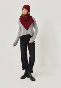 Piros bordázott sapka és illő oversized sál, szürke pulcsival, fekete, széles szárú nadrággal és fekete bokacsizmával párosítva. Egyszínű háttér.
