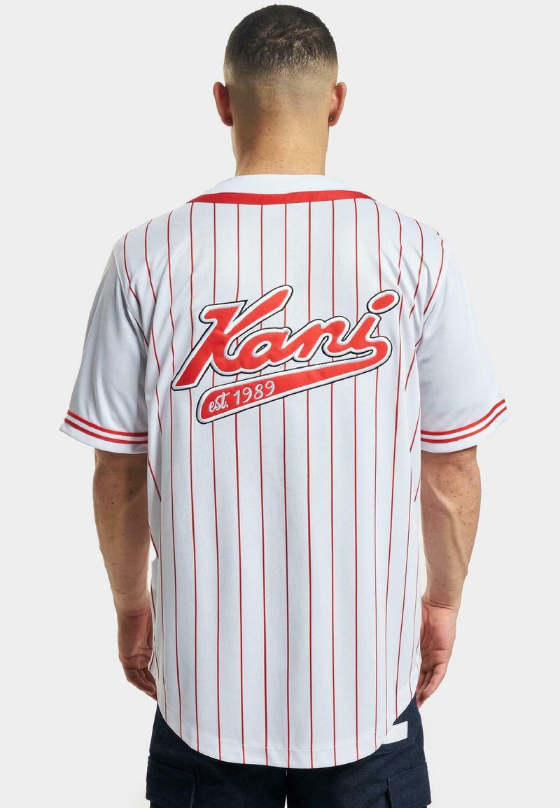 Tricou de baseball alb cu dungi roșii, având un mare logo roșu "Kani" pe spate și detaliul "est. 1989" dedesubt. Mâneci scurte.