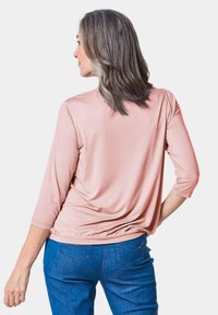 Hellrosa Bluse mit Dreiviertelärmeln und einem drapierten Rückendesign, kombiniert mit blauen Jeans, die ein dezentes gesticktes Detail an der Tasche aufweisen.