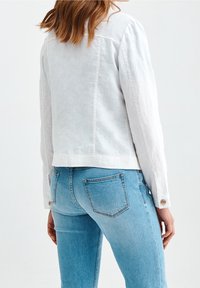Hvid linnedjakke med et cropped design, der har lange ærmer og knapdetaljer ved manchetterne, parret med lyseblå fitted jeans.