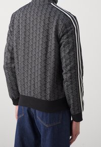 Czarna kurtka w wzory z geometrycznym designem, prążkowanymi mankietami i dołem oraz białymi paskami po bokach. Materiał mieszanki bawełny o teksturowanym wykończeniu.