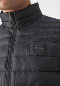 Svart vadderad jacka med hög krage, med horisontell söm, en framtida dragkedja och en liten brun "BOSS"-logotyp på vänster sida.